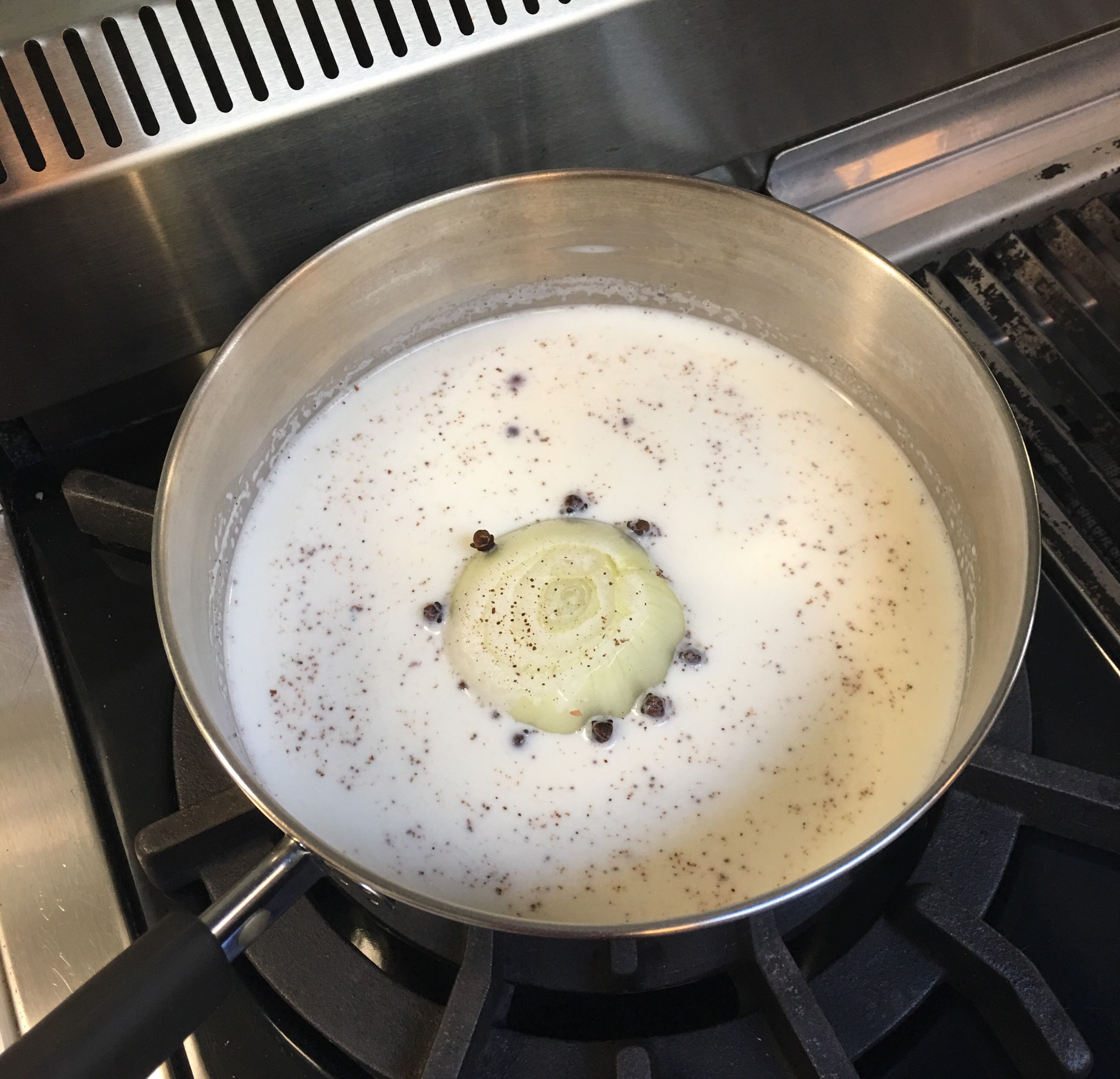 A pot on a stove containing milk and a onion with cloves stuck in the onion
